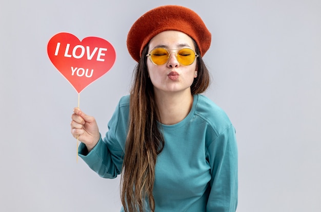 Mit geschlossenen Augen junges Mädchen am Valentinstag mit Hut mit Brille, das rotes Herz auf einem Stock hält, mit dem Text "Ich liebe dich", der Kussgeste isoliert auf weißem Hintergrund zeigt