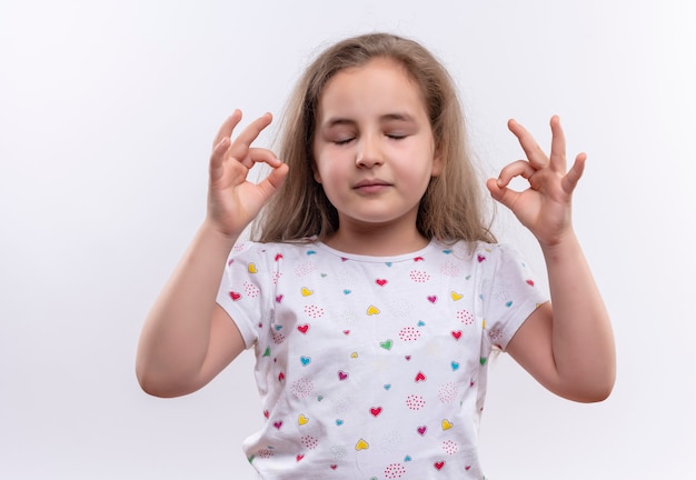 Mit geschlossenen Augen kleines Schulmädchen, das weißes T-Shirt trägt, das okey Geste auf isolierter weißer Wand zeigt