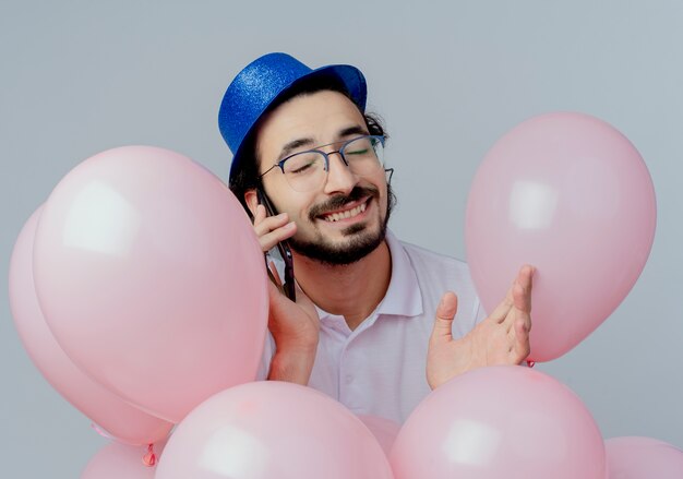 Mit geschlossenen Augen lächelnder gutaussehender Mann, der Brille und blauen Hut trägt, der unter Luftballons steht und am Telefon isoliert auf Weiß spricht