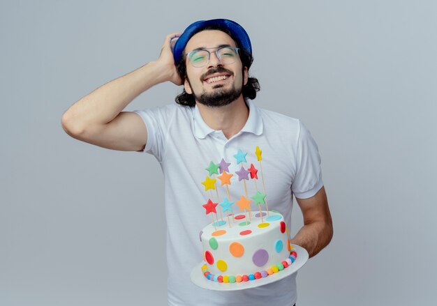 Mit geschlossenen Augen lächelnder gutaussehender Mann mit Brille und blauem Hut, der Kuchen hält und Hand auf den Kopf legt