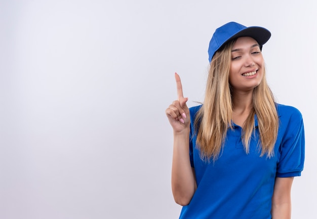 Mit geschlossenen Augen lächelndes junges Liefermädchen, das blaue Uniform und Kappe zeigt, zeigt oben auf weißer Wand mit Kopienraum