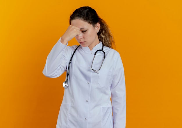 Mit geschlossenen Augen packte müde junge Ärztin in medizinischer Robe und Stethoskop die Nase isoliert auf orange Wand