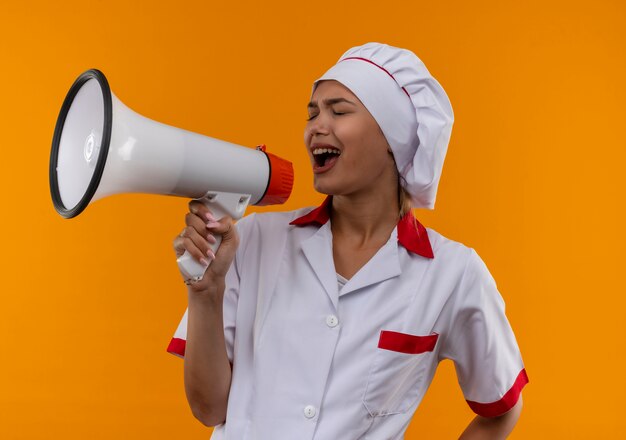 Mit geschlossenen Augen singt junge Kochfrau, die Kochuniform trägt, auf Lautsprecher auf lokalisiertem orangefarbenem Hintergrund