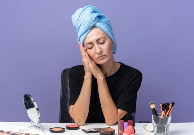 Mit geschlossenen Augen sitzt ein junges schönes Mädchen am Tisch mit Make-up-Tools und wischt sich die Haare im Handtuch ab und zeigt die Schlafgeste einzeln auf blauem Hintergrund