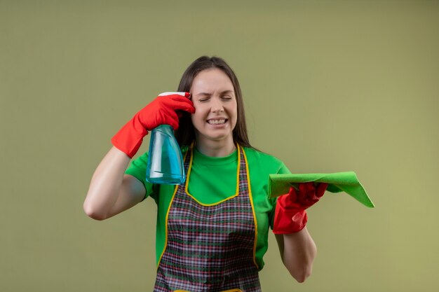 Mit geschlossenen Augen unzufrieden Reinigung junges Mädchen in Uniform in roten Handschuhen halten Reinigungsspray mit Lappen und Kratzer Wange auf isolierten grünen Wand