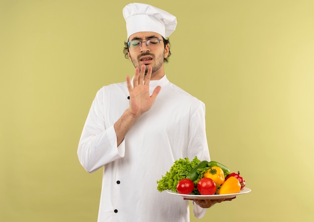 Mit geschlossenen Augen zimperlicher junger männlicher Koch, der Kochuniform und Gläser hält, die Gemüse auf Teller lokalisiert auf grüner Wand halten