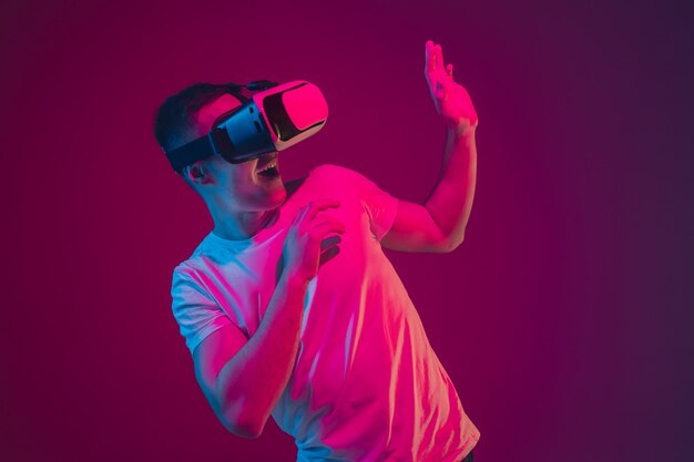 Mit VR spielen, filmen, fahren. Porträt des kaukasischen Mannes isoliert auf rosa-violetter Wand im Neonlicht. Männliches Model mit Geräten. Konzept der menschlichen Emotionen, Gesichtsausdruck, Verkauf, Anzeige.