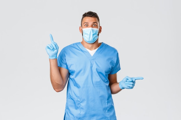 Mitarbeiter im Gesundheitswesen, Covid-19, Coronavirus und Viruskonzept. Aufgeregter männlicher Arzt, Krankenschwester in blauem Peeling und medizinischer Maske, persönliche Schutzausrüstung, Finger zur Seite zeigend, zwei tolle Neuigkeiten.