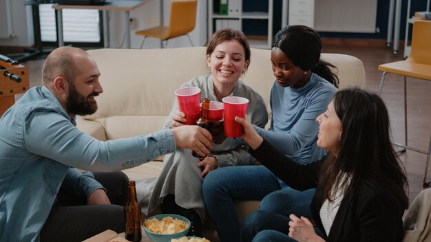 Mitarbeiter klirren mit Tassen und Flaschen, um die Freizeit nach der Arbeit zu genießen. Glückliche Kollegen jubeln mit alkoholischen Getränken, um Party zu feiern, Spaß und Unterhaltung nach Stunden im Büro zu haben.