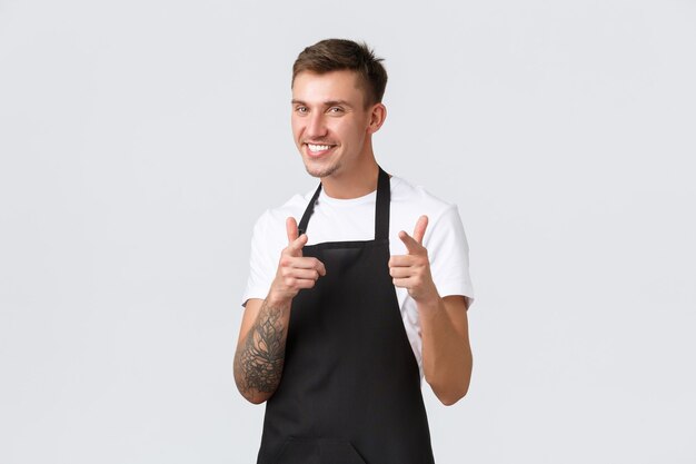 Mitarbeiter, Lebensmittelgeschäfte und Café-Konzept. Frecher hübscher blonder Kellner, Barista in schwarzer Schürze, breit lächelnd und mit den Fingern auf die Kamera zeigend, Gäste einladend, Kunden willkommen.