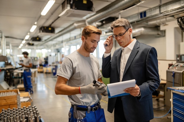 Mitte erwachsener Ingenieur und junger Arbeiter, die zusammenarbeiten, während sie Berichte in der Stahlfabrik analysieren