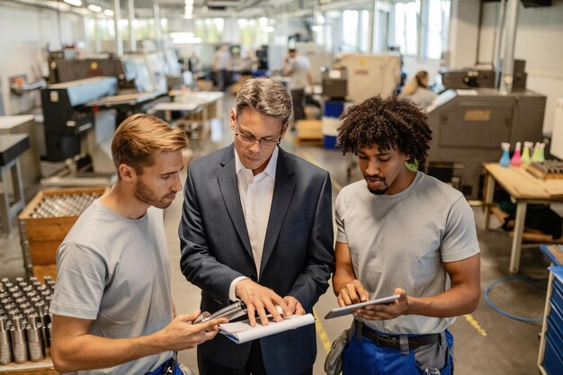 Mitte erwachsener Ingenieur und zwei Stahlarbeiter, die während ihrer Arbeit im Industriebau Qualitätsberichte durchgehen