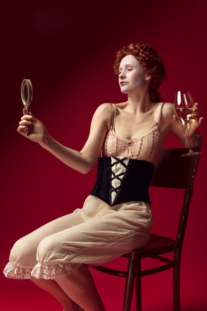 Mittelalterliche rothaarige junge Frau als Herzogin im schwarzen Korsett und in der Nachtwäsche, die auf roter Wand mit einem Spiegel und einem Glas Wein sitzen. Konzept des Vergleichs von Epochen, Moderne und Renaissance.