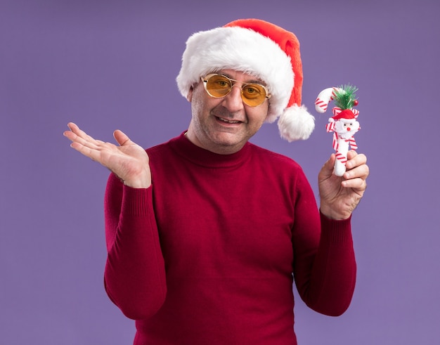 Mittelalterlicher Mann, der Weihnachtsweihnachtsmütze in den gelben Gläsern hält, die weihnachtliches Zuckerstangen-lächelndes Präsentieren mit Arm über lila Hintergrund halten