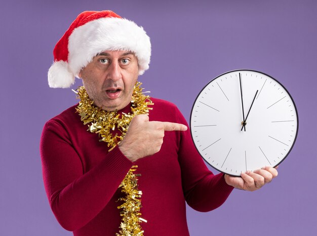 Mittelalterlicher Mann, der Weihnachtsweihnachtsmütze mit Lametta um den Hals trägt, der Uhr zeigt, die mit Zeigefinger darauf zeigt und besorgt über lila Hintergrund steht