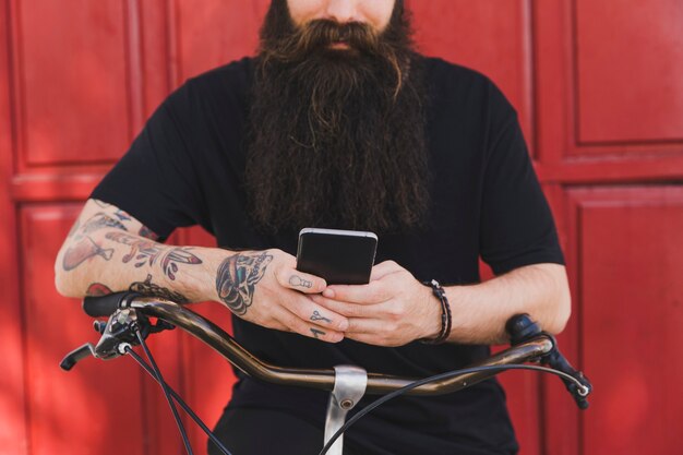 Mittelteil eines Mannes, der auf Fahrrad gegen rote Tür unter Verwendung des Handys sitzt