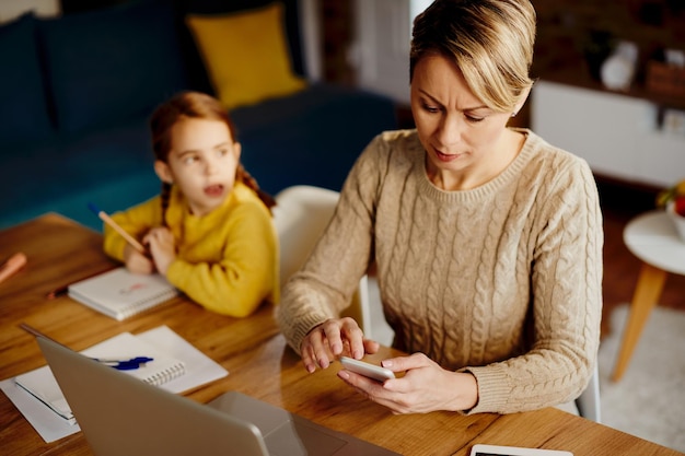 Mittlere erwachsene Mutter, die Smartphone verwendet, während sie zu Hause arbeitet