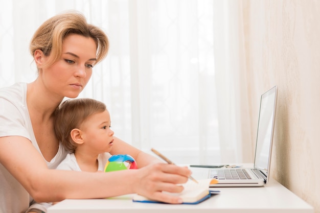 Kostenloses Foto mittlere schussfrau, die baby hält, das am schreibtisch arbeitet