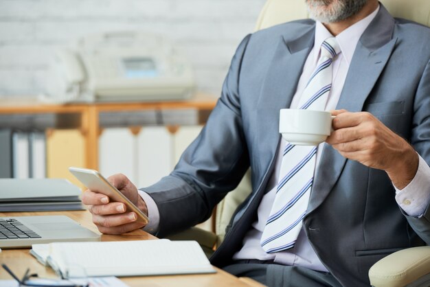 Mittlerer Abschnitt des trinkenden Kaffees des professionellen schauenden Angestellten und des Surfens des Netzes auf Smartphone