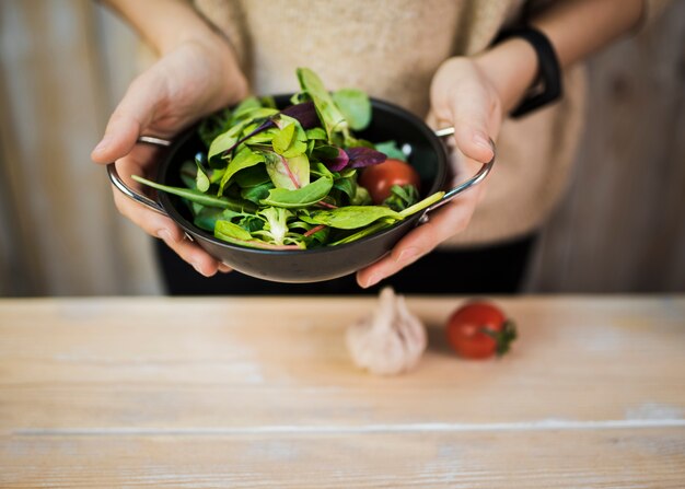 Mittlerer Abschnitt einer Frau, die frischen belaubten Gemüsesalat im Behälter über Holztisch mit Knoblauch und Tomate hält