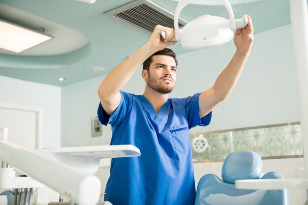 Mittlerer erwachsener männlicher Zahnarzt, der Beleuchtungsgeräte über dem Stuhl in der Zahnklinik anpasst