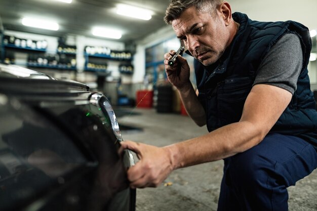 Kostenloses Foto mittlerer erwachsener mechaniker, der eine lampe verwendet, während er die vordere stoßstange eines autos in einer autowerkstatt untersucht