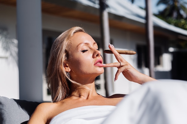 Mode-Außenporträt der nackten Frau sitzt auf Sofa, das sich mit Decke bedeckt, die Zigarre hält