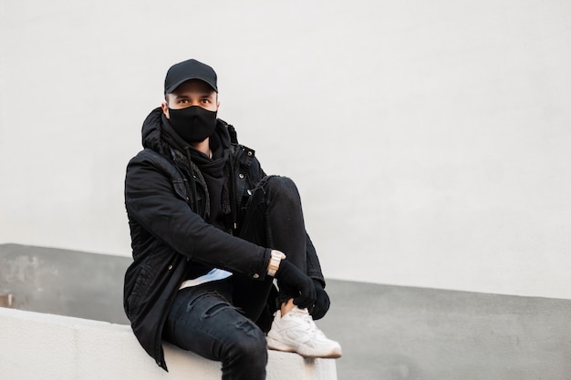 Mode-hipster-mann mit schutzmaske und schwarzer mütze in stylischer jacke und modischem hoodie mit turnschuhen sitzt auf der straße und bindet seine schnürsenkel.