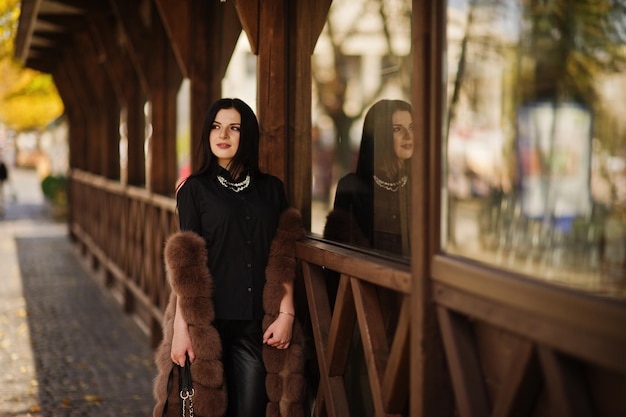 Mode-Outdoor-Foto einer hinreißenden sinnlichen Frau mit dunklem Haar in eleganter Kleidung und luxuriösem Pelzmantel vor Holzterrasse in der Herbststadt