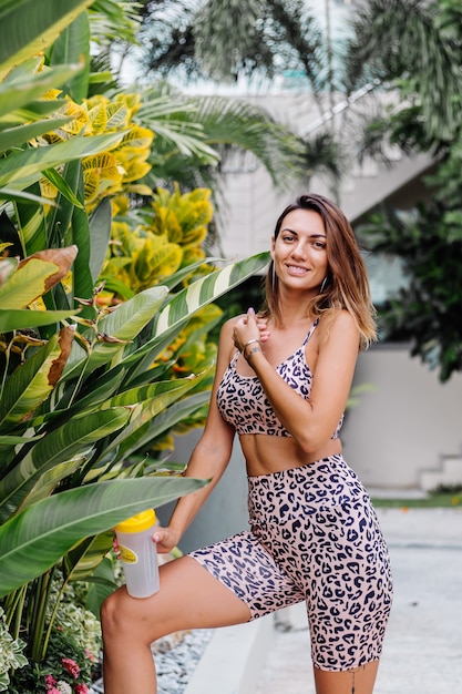 Mode stilvolle junge kaukasische Passform sportliche Frau in Leopard Cami Top und Biker Shorts außerhalb hält Protein-Shaker, Flasche Wasser, in der Hand