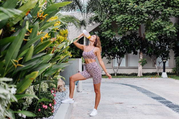 Mode stilvolle junge kaukasische Passform sportliche Frau in Leopard Cami Top und Biker Shorts außerhalb hält Protein-Shaker, Flasche Wasser, in der Hand