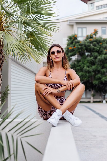 Mode stilvolle Passform gebräunte europäische Frau in Sonnenbrille, Leopard Cami Top und Biker Shorts, außerhalb Villa in der Nähe von Palme