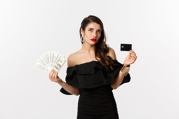 Mode- und Einkaufskonzept. Nachdenkliche Frau, die Kreditkarte und Dollar hält, nachdenkt und aufschaut, weißer Hintergrund