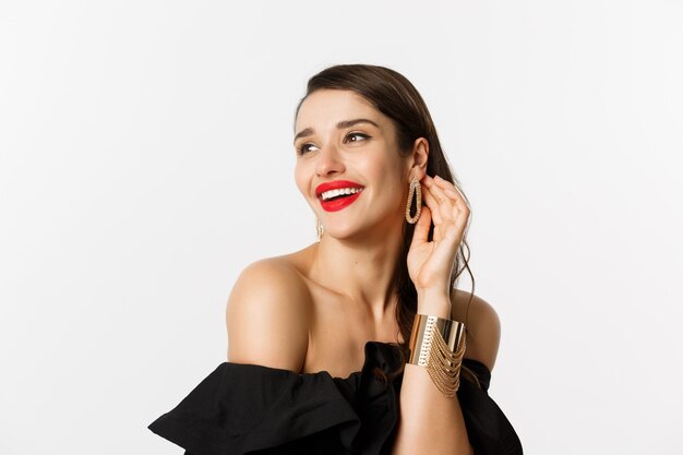 Mode- und Schönheitskonzept. Nahaufnahme einer eleganten brünetten Frau mit roten Lippen, schwarzem Kleid, kokett lachend und wegschauend, auf weißem Hintergrund stehend.