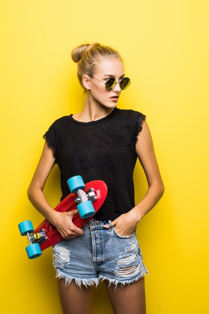 Mode ziemlich cooles Mädchen in der Sonnenbrille mit Skateboard über buntem gelbem Hintergrund