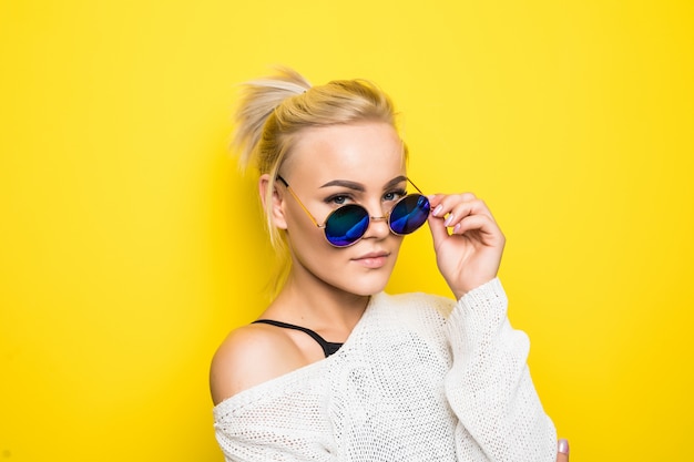 Modeblondes Mädchen im modernen weißen Pullover in der strahlend blauen Sonnenbrille posiert auf Gelb