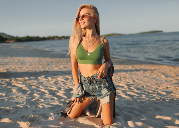 Modefoto der sexy blonden Frau im grünen Erntedach und in den Jeans, die auf tropischem Strand aufwerfen.