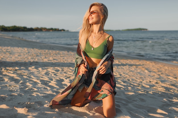 Modefoto der sexy blonden Frau im grünen Erntedach und in den Jeans, die auf tropischem Strand aufwerfen.