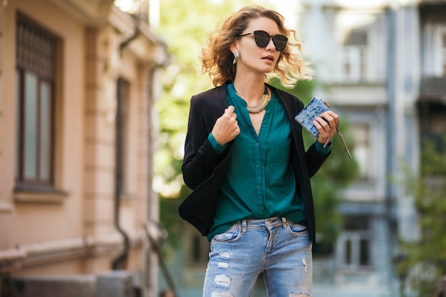 Modeporträt der jungen eleganten Frau, die in der Straße in der schwarzen Jacke, in der grünen Bluse, in den stilvollen Accessoires, im Halten der kleinen Geldbörse, im Tragen der Sonnenbrille, im Sommerstraßen-Modestil geht