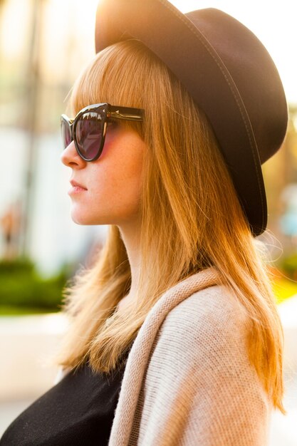 Modeporträt der jungen Hipsterfrau mit Hut und Sonnenbrille am Strand bei Sonnenuntergang, Retro-Stil Farbtöne Kalte Jahreszeit. Warme Kleidung.