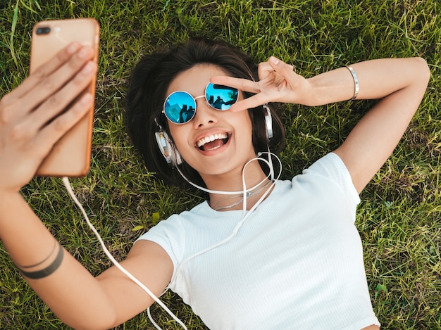 Modeporträt der jungen stilvollen Hipsterfrau, die auf dem Gras im Park liegt. Mädchen trägt trendiges Outfit. Lächelndes Modell, das Selfie macht. Frau, die Musik über Kopfhörer hört. Draufsicht