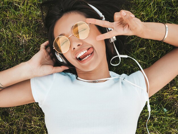 Modeporträt der jungen stilvollen Hipsterfrau, die auf dem Gras im Park liegt. Mädchen trägt trendiges Outfit. Lächelndes Modell genießen ihre Wochenenden. Frau, die Musik über Kopfhörer hört. Draufsicht