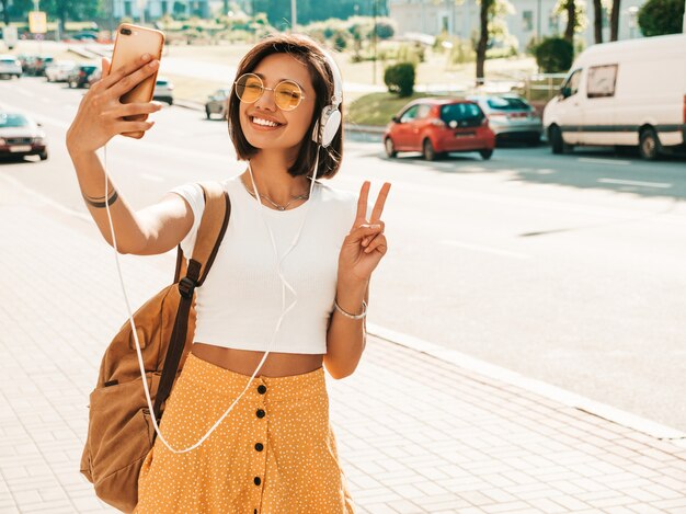 Modeporträt der jungen stilvollen Hipsterfrau, die in der Straße geht. Mädchen, das selfie macht. Lächelndes Modell genießen ihre Wochenenden mit Rucksack. Frau, die Musik über Kopfhörer hört