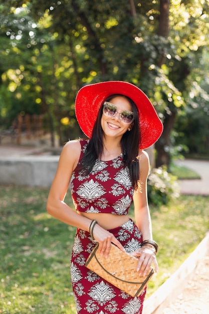 Modeporträt der lächelnden attraktiven stilvollen Frau, die im Park im Sommer-Outfit bedrucktes Kleid geht und trendige Accessoires, Geldbörse, Sonnenbrille, roten Hut trägt, im Urlaub entspannend