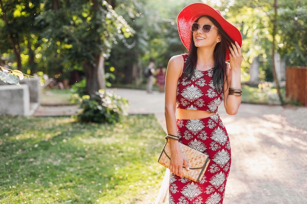 Modeporträt der lächelnden attraktiven stilvollen Frau, die im Park im Sommer-Outfit bedrucktes Kleid geht und trendige Accessoires, Geldbörse, Sonnenbrille, roten Hut trägt, im Urlaub entspannend