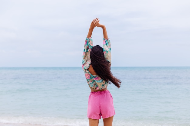Modeporträt der stilvollen Frau im bunten Druck-Langarmoberteil und in den rosa Shorts am Strand, tropischer Hintergrund.