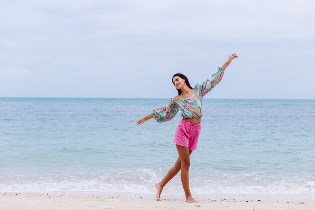 Modeporträt der stilvollen Frau im bunten Druck-Langarmoberteil und in den rosa Shorts am Strand, tropischer Hintergrund.