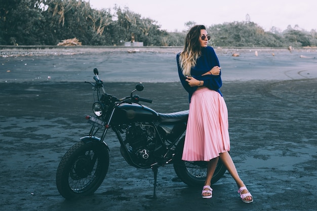 Modeporträt der stilvollen Frau in der Sonnenbrille, im blauen Pullover und im rosa Rock, steht durch schwarzes Fahrrad auf schwarzem Sandstrand.