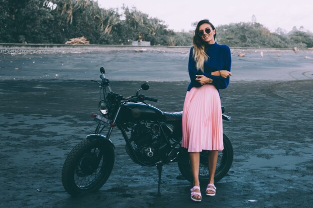 Modeporträt der stilvollen Frau in der Sonnenbrille, im blauen Pullover und im rosa Rock, steht durch schwarzes Fahrrad auf schwarzem Sandstrand.