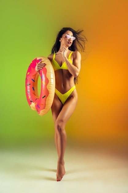 Modeporträt einer jungen, fitten und sportlichen Frau mit Gummikrapfen in stylischer gelber Badebekleidung an einer Wand mit Farbverlauf, perfekter Körper für den Sommer bereit